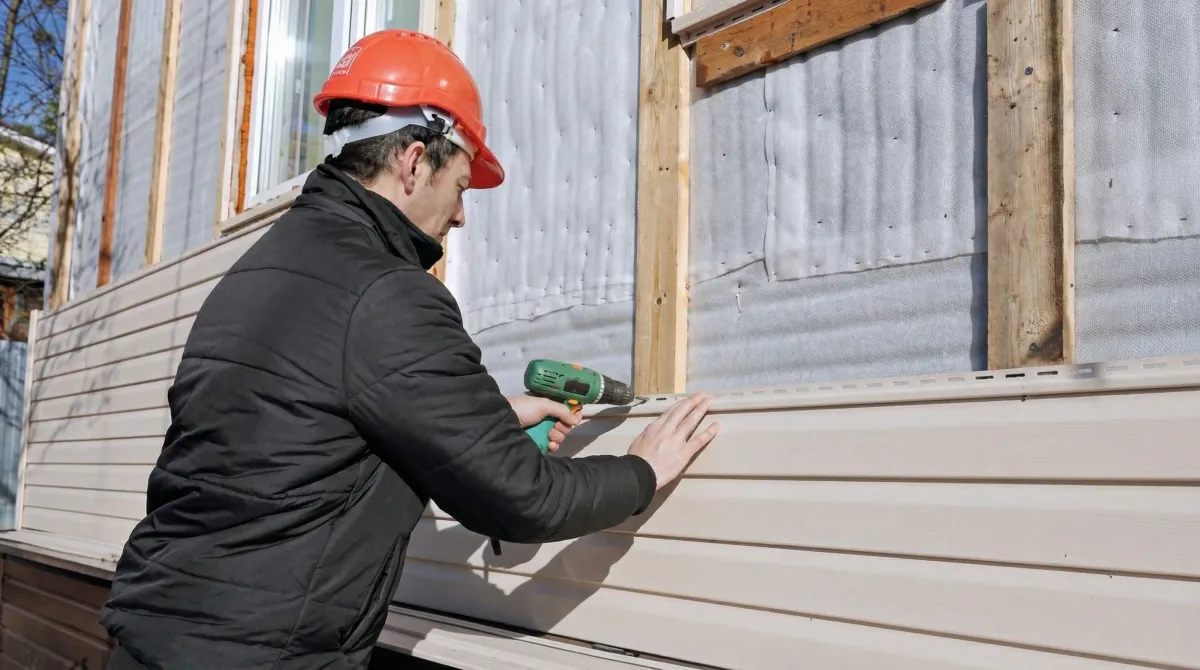 Siding replacement professional using a power drill to install new tan vinyl siding on a home exterior in Riverside, CA